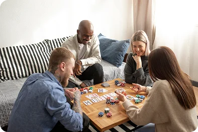 Vier lachende vrienden die gezellig samen een kaartspel met fiches spelen aan een houten salontafel in de woonkamer.